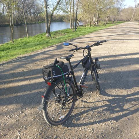 A block bicycle with a tail bag on a rear rack on a gravel road alongside a river to scene left. A setting sun from the left rear creates shadows in from trees in front of the bicycle from left to right. A strip of green grass is paralle to the road along the left side