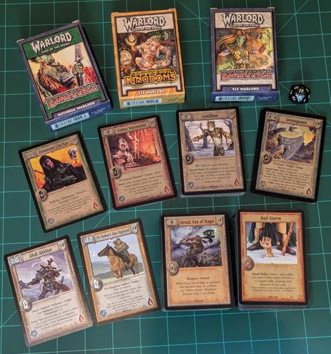 A small selection of Warlord Saga of the Storm cards. There is one of each faction, item and action card. At the top are some of the original starter decks and a d20 die.
