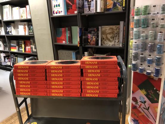 A trolley with three piles of copies of a bright red book with yellow and black lettering on the spine. Bookshelves are behind it, left and centre. On the right are miscellaneous stationery supplies.