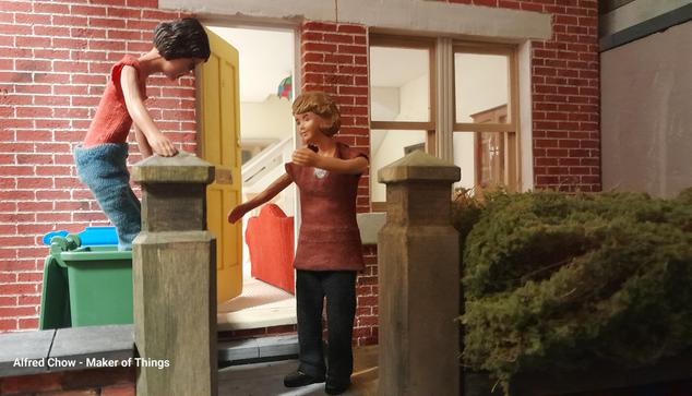 A 1:12 scale diorama of the outside of Daisy's house, looking at the house front. The house us red brick with a yellow front door, opened, and a double sash window to the right. There is a small front garden with an overgrown hedge and stone gateposts. Behind the gateposts are two wheelie bins, one blue and one green. The green bin has its lid open.

Harriet, a Caucasian woman with dark hair and wearing a red tee shirt and blue jeans, is standing in the green bin trying to compress the rubbish down, she is holding onto the top of a gatepost to steady herself.

Daisy, a Caucasian woman with fair hair and wearing a burgundy top and black trousers, is standing on the path with her arms out in case Harriet falls.

Harriet: If I can just…. Bounce, bounce, bounce....
Daisy: Be careful!
Harriet: We only have a bit more rubbish to get in here.
Daisy: But that’s no good if it is too jammed in to tip out!
Harriet: Oh, yeah. That’s a good point.
Daisy: I'll help you get down.