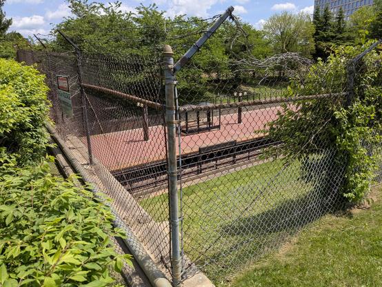 View of the corner of a rusty chain link fence, in front of a grassy embankment that drops down to the red-bricked boarding platform for the North Bethesda Metro train station. Bright green shrubbery forces itself up against and all over the fencing, with dimmer trees in shadow in the background. Vivid green foliage contrasts with the angular reds of the fence and platform.