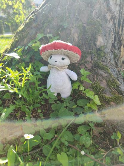 A crocheted doll resembling a mushroom stands against the base of a large tree. The doll has a white body, black eyes, and a red and beige mushroom cap on its head. The ground around the doll is covered in green leaves and small plants, with sunlight filtering through the foliage.