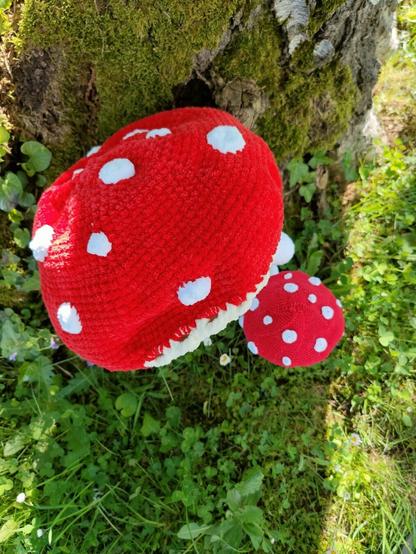 Two crochet mushroom dolls, one large and one small, with only their red, white-spotted hats visible. The hats rest near a moss-covered tree trunk, surrounded by green grass and small plants.