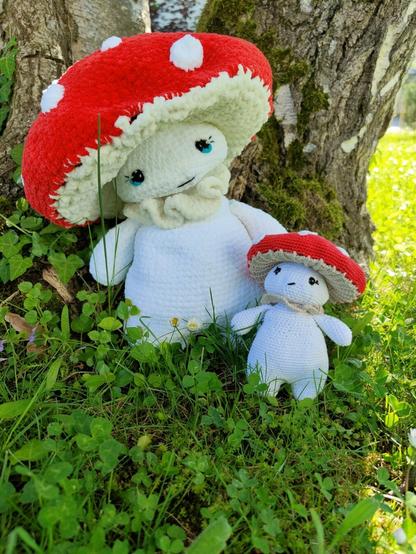 Two crochet mushroom dolls with red caps and white spots sit at the base of a tree. The larger doll has blue eyes and a cheerful smile, while the smaller one has black eyes and a neutral expression. They are surrounded by green grass and small plants, with sunlight filtering through the foliage.