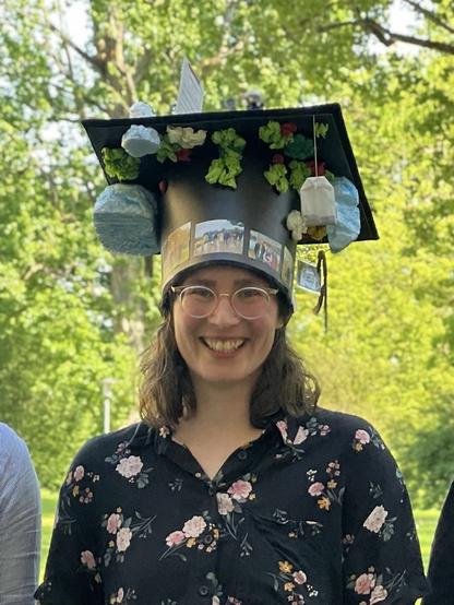 For a PhD defence in Germany, it is good tradition that all lab members collaborate in crafting a black hat out of cardboard, mimicking a traditional graduation hat with a square surface on top. The hat is then typically decorated with stuff related to the PhD thesis or the PhD candidate, in this case several pictures, bryozoans, icebergs, a tea bag, mathematical symbols and a lot more. The picture shows Dr. Franziska Koch, a white woman with dark hair and glasses, proudly wearing her new hat and smiling into the camera. The picture was taken outside, the background is bright green and blurry.