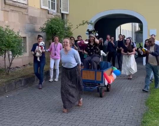 Another German tradition requests that the former student takes her seat on a wagon that is then pulled across the campus in order to present the freshly awarded doctor to the academic world. The wagon in the picture is nicely decorated with balloons and pulled by the former supervisor (me, a white woman with blond hair, wearing a long black skirt and a blue blouse). To draw even more attention of the academic world, the attending colleagues make A LOT of noise by cheering and clapping.