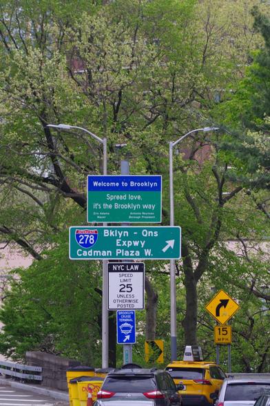 The signpost at the Cadman Plaza offramp of the Brooklyn end of the Brooklyn bridge. On top is a "welcome to Brooklyn" sign with slogan "Spread love, it's the Brooklyn way", followed by other traffic signs below.