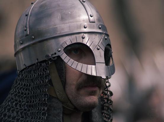 A close-up photo of a man wearing what appears to be medieval armor, specifically designed for combat. Below the helmet, part of a chainmail coif (a garment made up of individual metal rings linked together) is visible around the neck area. The design suggests that this could be from a historical reenactment. The man's face is partially obscured by the helmet, but his mustache adds a touch of personality to his appearance. He looks very seriously to the right side of the frame.
