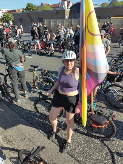 Mirah wearing a light purple crop top, black shorts, and a bike helmet standing in front of her bike on a sunny Autobahn. There's a full size progress pride flag on a pole attached to her bike