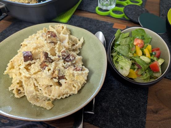 Links ein grüner tiefer Teller mit Mafaldine Carbonara, rechts daneben ein Schälchen mit Salat aus Romana, Tomate, gelber Paprika, Gurke & Schnittlauch. Im Hintergrund sind die Ecke einer Pfanne und ein grüner Untersetzer zu sehen