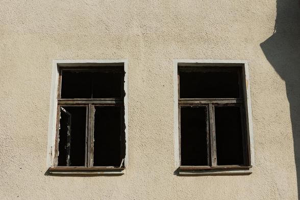 Photo of grayish wall with two windows with wooden frames but without glass. Bright sunlight on the wall and black holes of windows make very dramatic contrast.
