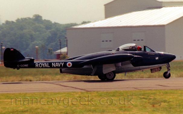 Side view of a black, single engined military jet fighter taxiing from left to right along a grey runway.
The plane is basically a large black engine pod with a white belly, with twin black tail booms stretching backwards from the wing root, carrying the small twin tails.
The cockpit canopy at the front of the plane is open, slid back along the top of the fuselage, with the bright red helmet of the pilot clearly visible in the gap.
The tail booms has white "Rioyal Navy" titles, just aft of a blue, white, and red roundel.
The registration "G-GONE" is on the end of the boom in white, under the tail.
Green grass fills the foreground, with a large grey building dominating the frame on the right, and trees on the left in the background, under bright but hazy skies.