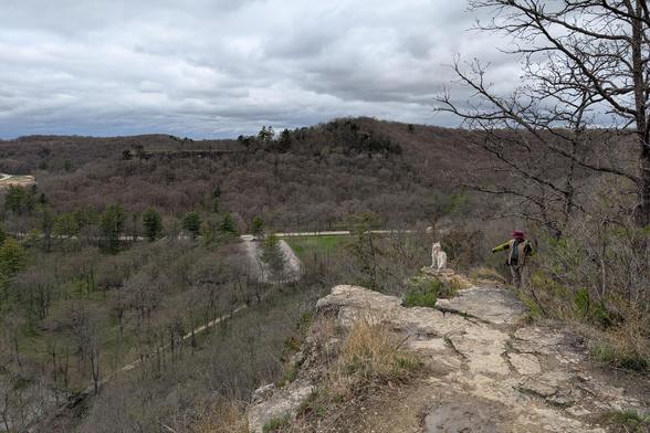 Jeff continues to photograph Zeena on the bluff, while the photographer has passed and is looking down from an even higher overlook on the edge of the bluff. From here, you can see the valley below, and bluffs of similar height on the far side. A road cuts through the valley, and a park parking lot can be seen between trees below. The flat, textured cloud layer undulates over the far hills.