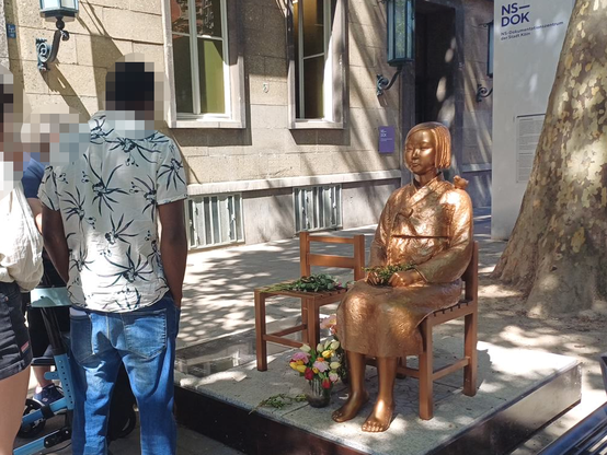 Eine Gruppe Menschen steht vor einer Skulptur einer sitzenden "Trostfrau" (Zwangsprostituierte des japanischen Militärs) vor dem El-De-Haus