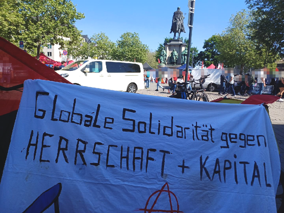Banner "Globale Solidarität gegen Herrschaft und Kapital" (A-Forum Köln), im Hintergrund das DGB-Straßenfest beim Reiterdenkmal auf dem Heumarkt