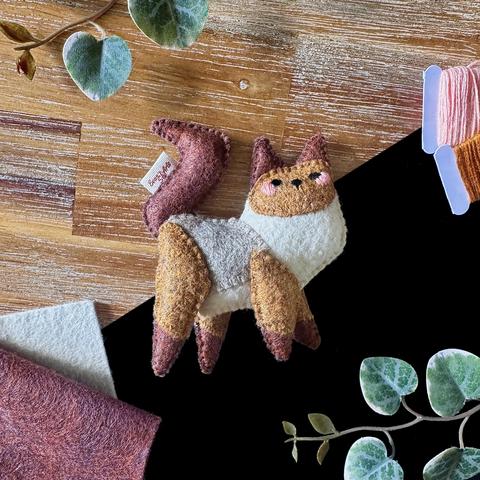 A handmade Birman cat felt plushie photographed on a black and wooden background, surrounded with decorative plants, felt and threads. 
The cat is standing sideways but is facing the viewer. It has dark brown ears, feet and tail and a lighter brown face and legs. It’s back is a soft grey. Small squinty eye, a small nose and a soft pink blush are embroidered on it’s face.