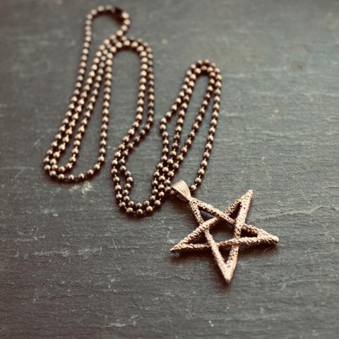 A photo of a vintage pewter pentagram pendant necklace from the 1980s on a dark grey slate background. The pentagram pendant is large, rustic/textured, and 3-dimensional. It hangs from a blackened 30" stainless steel ball chain.