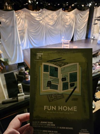 Fun Home playbill held in a hand of a person sitting in a balcony of a theater, viewing the stage in the background.