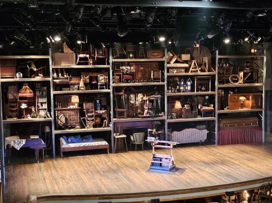 Stage for the musical Fun Home. There are racks of furniture filling from stage to rafters at the back of the stage. In the center of the stage is an artist's easel.