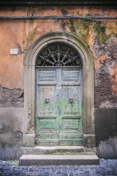 In Viterbo Italy, an old wooden door with peeling green and blue paint sits in a stone archway, surrounded by a weathered orange and gray concrete wall. Intricate ironwork above the door adds a touch of elegance to the rustic facade.