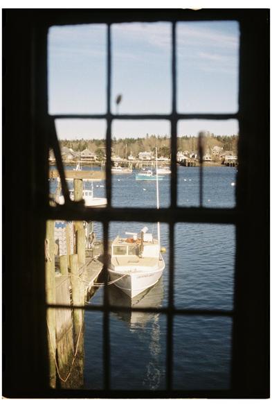 A 35mm film photo taken through a closed window. The window muntins are out of focus in the foreground and the subject of the photo, a lobster boat tied up at a float, is in focus in the background.