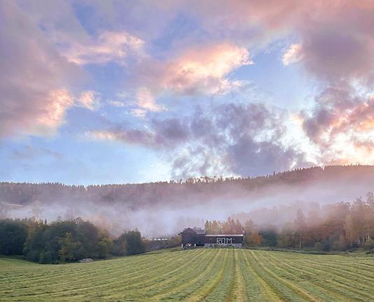 The image shows a picturesque rural landscape during what appears to be early morning, as indicated by the soft light and mist. There's a field in the foreground with visible traces from agricultural activity. The field leads up to a building that looks like a farm or house, with the letters "ROM" visible on its side. Beyond the building, a layer of mist partially obscures a dense forest covering rolling hills. Above, the sky is filled with puffy clouds tinged pink and orange by the sunrise, suggesting a peaceful and serene start to the day.