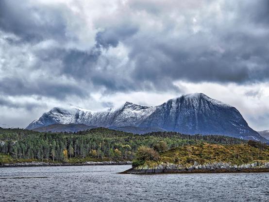 The image shows a dramatic landscape with a rugged mountain range covered partially in snow in the background. There's a large body of water in the foreground, possibly a lake or a fjord, bordered by a landmass with dense greenery and scattered trees. A small island or peninsula is prominently shown in the foreground featuring grass and trees. The sky is overcast with heavy clouds, suggesting a moody or chilly atmosphere. The overall setting appears to be in a cold, northern climate, possibly in Scandinavia or a similar region.