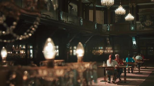 A dark room, dimly lit by candelabras and overhead lights, and with a balcony overlooking the action. There are three light wells where games of chess are going on, casting shadows away from the windows.