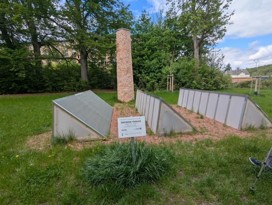 Foto der Installation. Zu sehen ist ein kleiner ca 4m hoher Schornstein Auf einer Wiese. Davor drei schrägdächer, die wie eine Industrieanlage aussehen, aber auch nur auf der Wiese stehen. Das schild der Installation zeigt nur auf, dass es iskander yediler heißt und Teil des purple path ist. Kein qr code, nix. Von braille Schrift auch keine spur