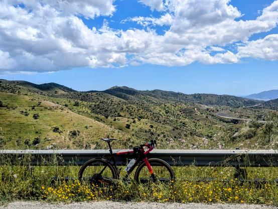 My bike against the guardrail on the road, with hills in the background, mostly blue sky with some non menacing clouds.