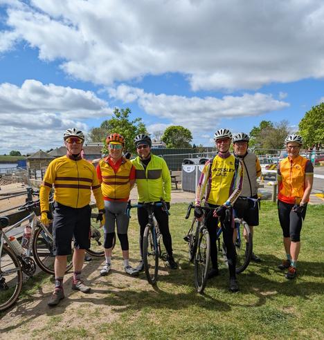 Six cyclists pose for a photo. They are wearing tops that are either yellow, green or grey. They are stood on grass and the sky is partially cloudy.