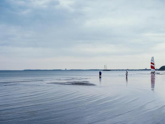 The image shows a calm beach scene with a few people wading in shallow water, a red-and-white striped sailboat near the shore, and a distant pier stretching out into a tranquil sea under a cloudy sky.