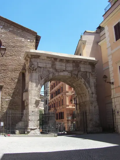 The image shows an architectural structure known as an arch, likely a historical monument. It appears to be made of stone and is partially surrounded by modern buildings. The sky above is clear and blue, indicating a bright day. Some fencing is visible around the arch, possibly for preservation or safety purposes.

Image Credits: Wikimedia / FlagUploader (User:Panairjdde) / CC SA 1.0