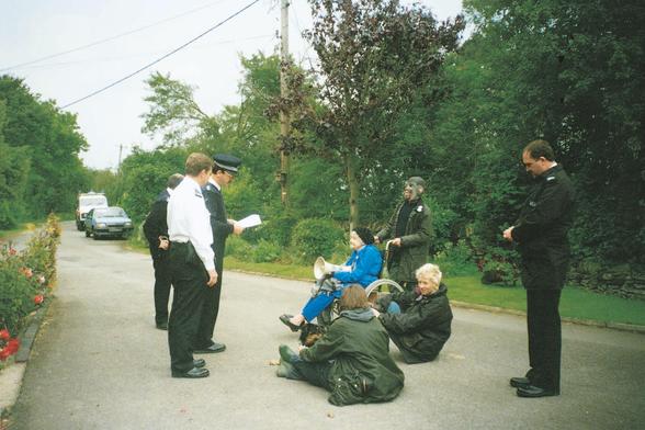 Fotografía: cuatro policías piden la documentación a cuatro activistas que protestan en un camino cercano al criadero de gatos destinados a la experimentación de Hill Grove. Esto ocurrió en Reino Unido a finales de los 90. Dos activistas están sentadas en el suelo, con un perrete. Otra activista, la que lleva el megáfono, es una anciana muy adorable sentada en una silla de ruedas. Y de pie sujetando la silla un o una activista con una máscara de mono.