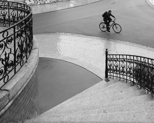 Der Blick führt eine geschwungene steinerne Treppe hinunter zu einer ebenso geschwungenen T-Kreuzung, die ein Radfahrer von links nach rechts überquert.
Das Foto ist schwarz-weiß. Der Mann auf dem Fahrrad ist bewegungsunscharf abgebildet. Links ist eine Art Veranda – von einem runden verzierten Metallgeländer umgeben. Der Betrachter scheint in gleicher Höhe am Beginn der Treppe zu stehen. Rechts im Vordergrund ist die nach unten führende Spindeltreppe, rechts mit einem Geländer gesichert. Davor läuft ein Gehweg entlang, geschwungen und glänzend vor Nässe. Zwei weitere geschwungene Gehwege entlang der Kreuzung sind im Hintergrund. Die Treppe führt auf eine kleine asphaltierte Fläche vor dem Gehweg. Der sichtbare Teil hat die Form eines Tortenstücks, so wie auch der Teil der Treppe. Geländer, Mauer, Fahrbahn- und Gehwegränder – alles besteht aus Kreisbögen. Im Kontrast zu den statischen, architektonischen Elementen ist der Radfahrer in Bewegung. Aber auch im Fahrrad wiederholen sich Kreise und Dreiecke.

The view leads down a curved stone staircase to an equally curved T-junction, which a cyclist is crossing from left to right.
The photo is black and white. The man on the bicycle is shown motion-blurred. On the left is a kind of veranda - surrounded by a round, decorated metal railing. The viewer appears to be standing at the same height at the beginning of the stairs. On the right in the foreground is the spiral staircase leading downwards, secured on the right by a railing. A