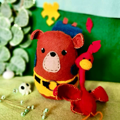 A picture of a felt plushie in front of a green background with decorative plants, a small skull, a jigsaw piece, and a feather to decorate. The plushie is of a simplistic brown bear; basically a potato with ears. It has a yellow short with a black belt and is wearing a small felt blue backpack. Next to the bear is sitting a red bird with a big red beak and a long thin neck.