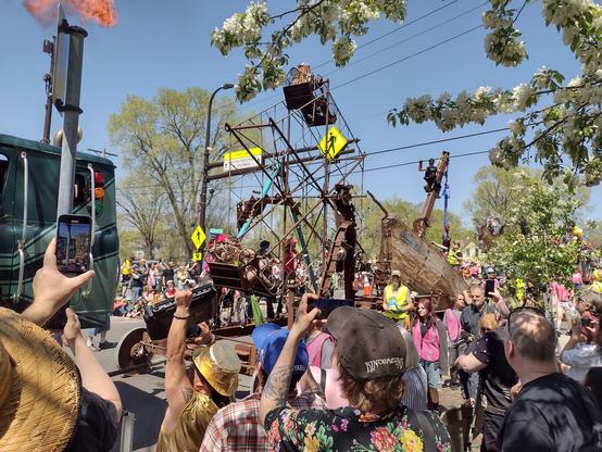 A large 2 story high ferris wheel parade float in a street parade framed by tree blossoms. .  The vehicle towing the float is visible to photo left with a large gout of flame visible top left.
