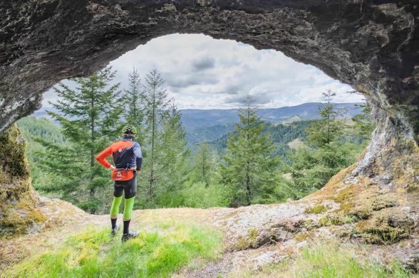 A hiker stands at the entrance of a natural rock formation, looking out over a mountainous landscape filled with trees and rolling hills under a partly cloudy sky. The hiker is wearing bright clothing and appears to be enjoying the view