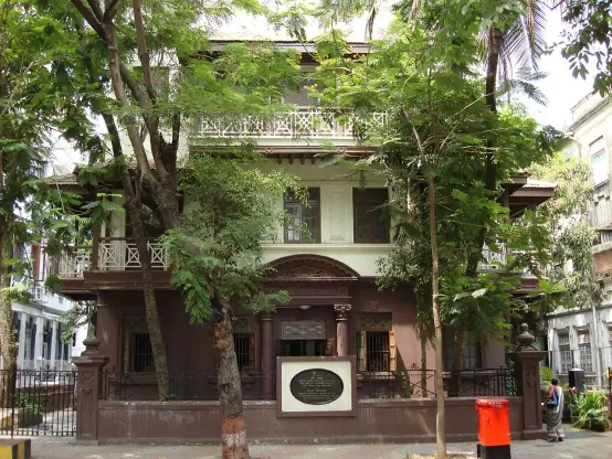 The image shows a large, two-story building surrounded by trees. The architecture features ornate details, including balconies and a decorative facade. There is a sign in front of the building, likely with information about its significance. The area appears to be urban, with other buildings visible nearby.

Image Credits: Wikimedia / Jorge Láscar / CC BY-SA 2.0