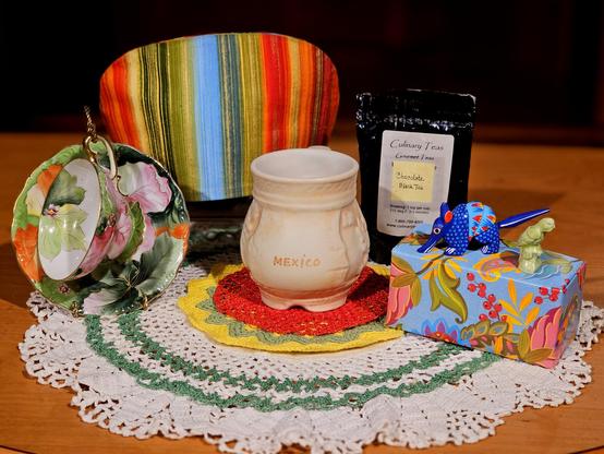 Tea tableau to celebrate Mexican-American heritage on Cinco de Mayo with bright, ruffled textiles and colorful china, a Mexican pottery mug, a wooden carving of an armadillo, a green ceramic parrot, and chocolate flavored black loose-leaf tea.
