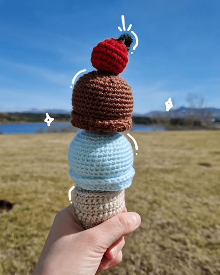 A hand holding a crocheted ice cream cone with two scoops—light blue, brown, and a red cherry on top—against a scenic outdoor background with a grassy field, blue lake, and mountains under a clear blue sky. White doodles highlight the ice cream scoops.