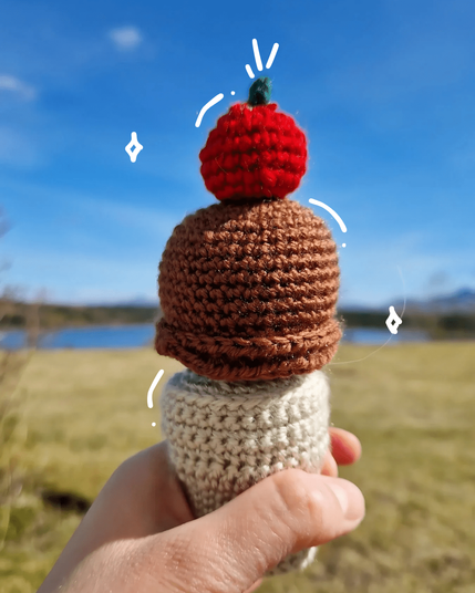 A hand holding a crocheted ice cream cone with one brown scoop and a red cherry on top—against a scenic outdoor background with a grassy field, blue lake, and mountains under a clear blue sky. White doodles highlight the ice cream scoops.