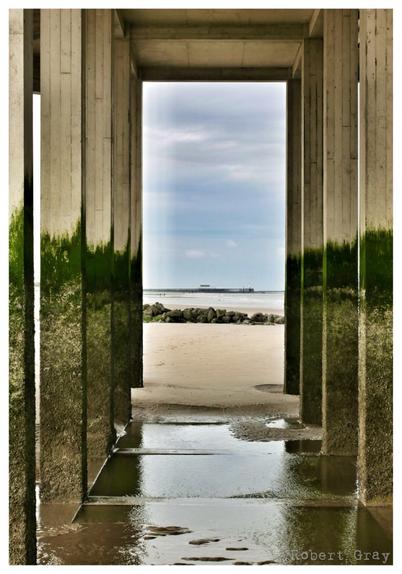 A view of the distant coast taken through the algae-coated concrete supports of a pier.