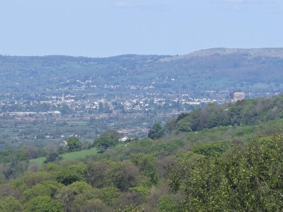 A picture of Cheltenham town from a hill. You can see various hills in the background. Various buildings are displayed, including the Eagle Tower (tallest building) and a church looking structure.