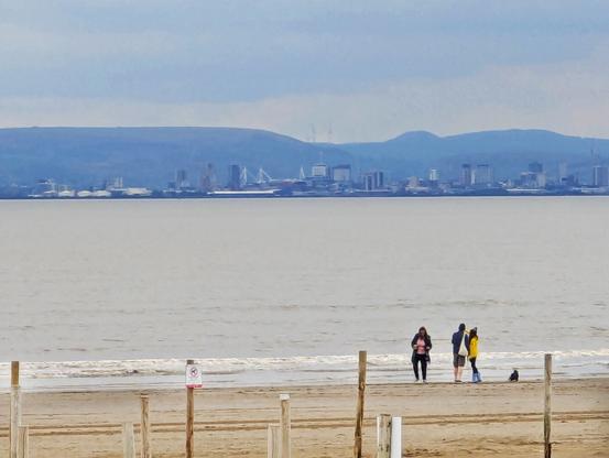Cardiff skyline from Weston-super-mare in North Somerset. The sea and beach are visible, alongside hills north of Cardiff. A group of people are on the beach with a dog, fencing and a sign saying no dogs is also visible.