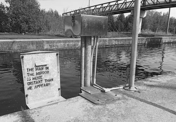 A steel electrical box next to a canal has the words "The man in the mirror is more distant than he appears" stenciled on it. Other electrical boxes are seen as well as a footbridge crossing the water on the top right of the image. Trees can be seen on the other side of the canal in this black and white image.