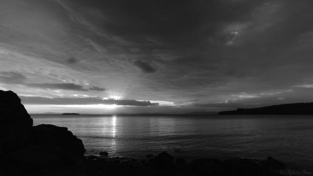 An ominous, dark cloud fills most of the sky, as the sun sets in the distance across a large body of glassy rippled waters.  A starburst surrounds the sun as it peeks under a cloud, rays of light scattered above in a fan-like splay, lighting the underside of the cloud layer above.  Foreshore rocks and nearby headlands lay in silhouette, contrasting heavily against the bright sunlight reflected in the sea.  Photographed in black and white using in-camera features.