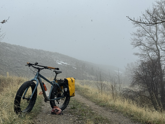 A blue fat bike with yellow pan is parked on a gravel 2 track. Fog and blowing snow obscure the hill behind the bike..