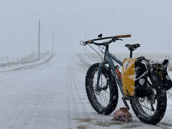A blue snow covered fat bike with yellow pans on a snow covered gravel road. Blowing snow is working its way toward a white out..