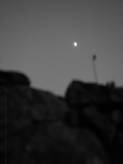 The image is blurry. It appears to show a bright spot in the sky, possibly a celestial object like the moon or a bright planet, above a dark, indistinct foreground that could be rocks.